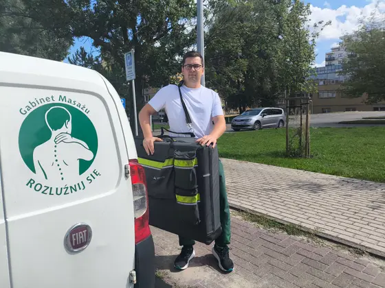 Masażysta wyjmuje z auta składany stół i torby ze sprzętem do masażu